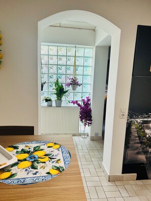 Een kamer met betegelde vloer, een witte radiator, een tafel met tafelkleed met patroon en een raam met uitzicht op een stadsbeeld.