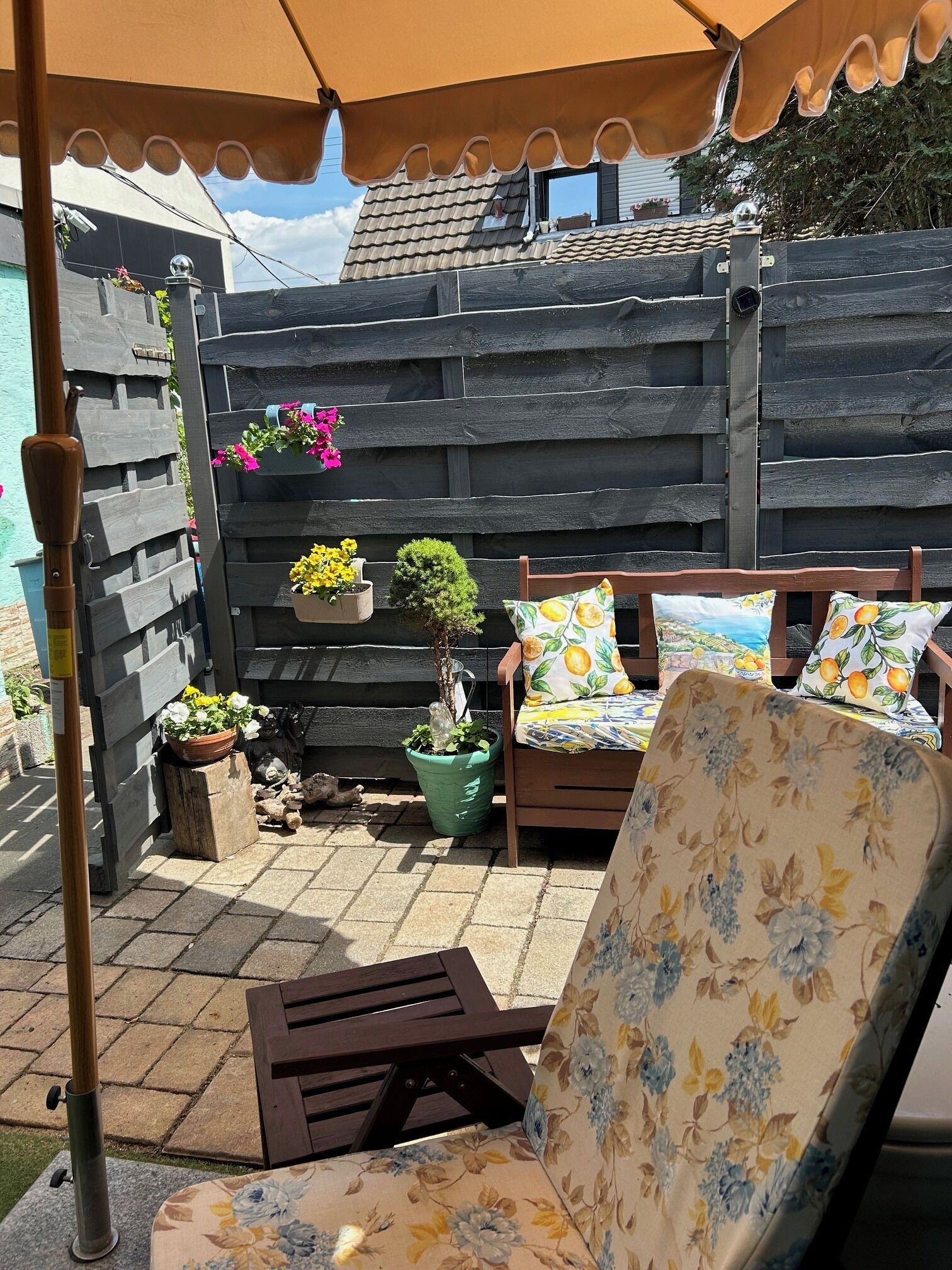 A patio with a wooden fence, a wooden chair with floral cushions, a small table, and potted plants.