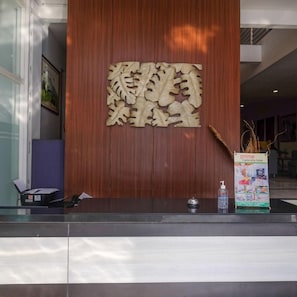 A reception desk with a wooden panel featuring carved text, a glass partition, a framed picture on the wall, and a hotel sign. - Prime Cailendra Hotel Tamansiswa Yogyakarta (Yogyakarta)