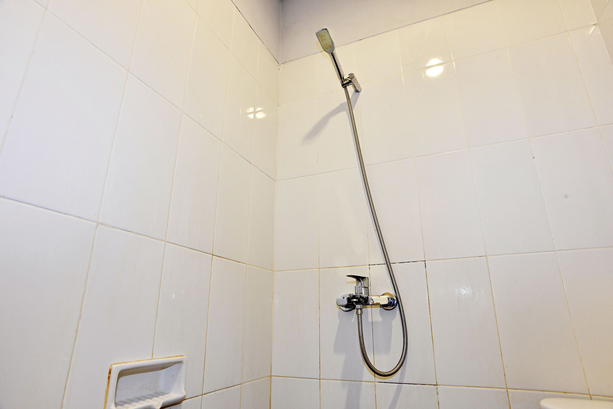 Une petite salle de bain d’angle avec des murs recouverts de carreaux blancs, une douche avec un pommeau de douche amovible et un petit lavabo blanc.