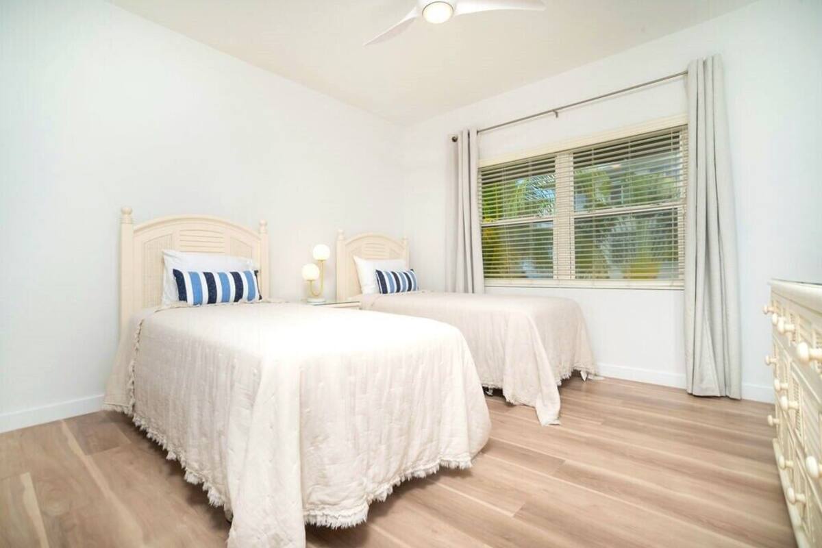 Quarto com duas camas, um ventilador de teto, janela com persianas e piso de madeira.