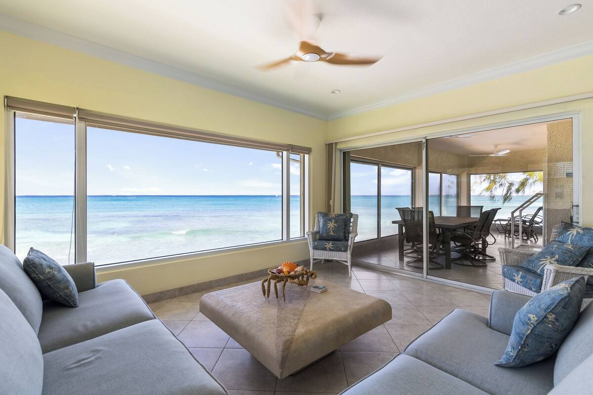Quarto espaçoso com grande janela com vista para o oceano, ventilador de teto e área de jantar com mesas ao ar livre.