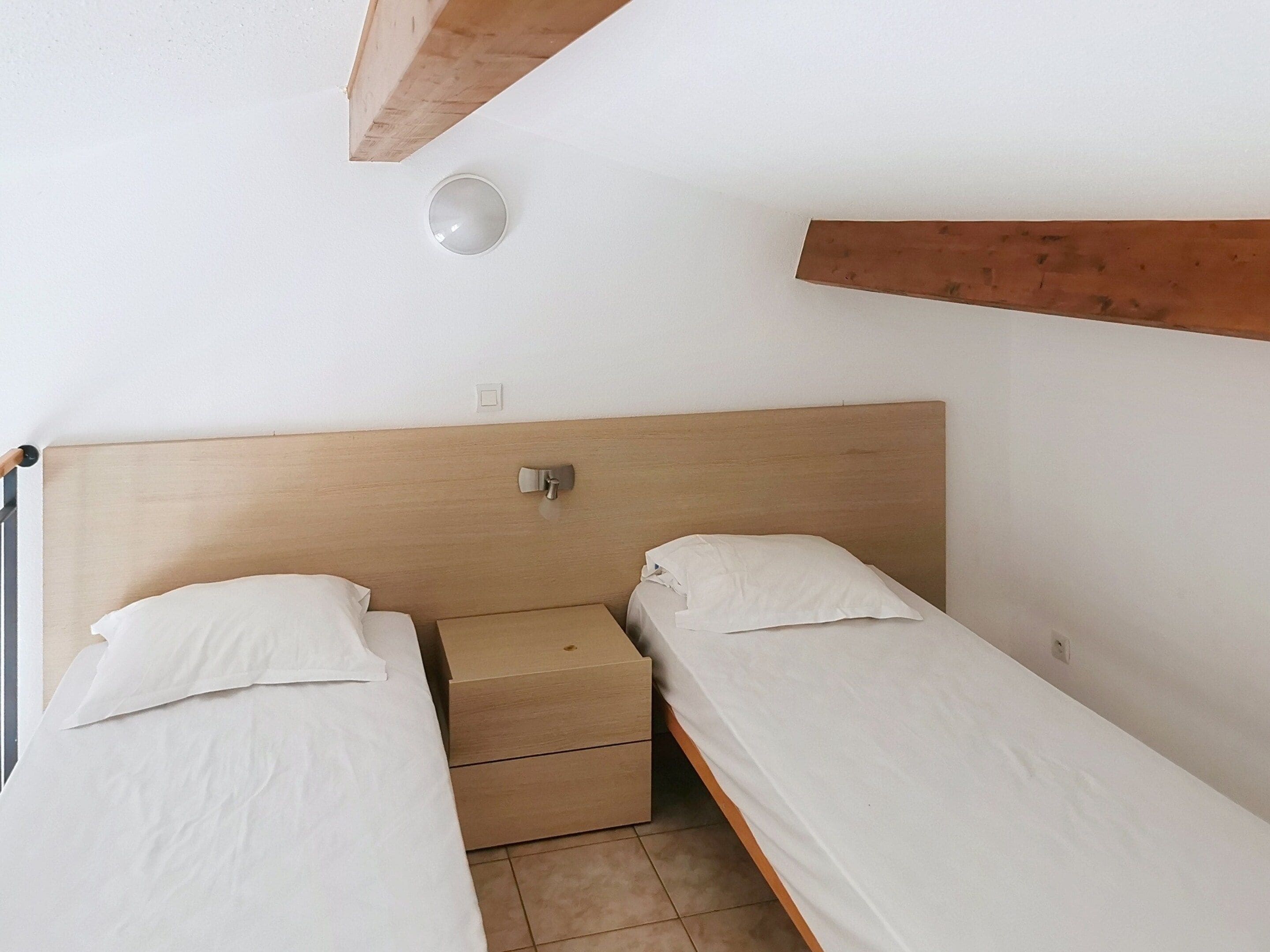 Une petite chambre simple, avec deux lits simples, une tête de lit en bois, une table de chevet et un luminaire fixé au mur.