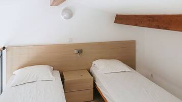 A small, simple room with two single beds, a wooden headboard, a bedside table, and a wall-mounted light fixture.