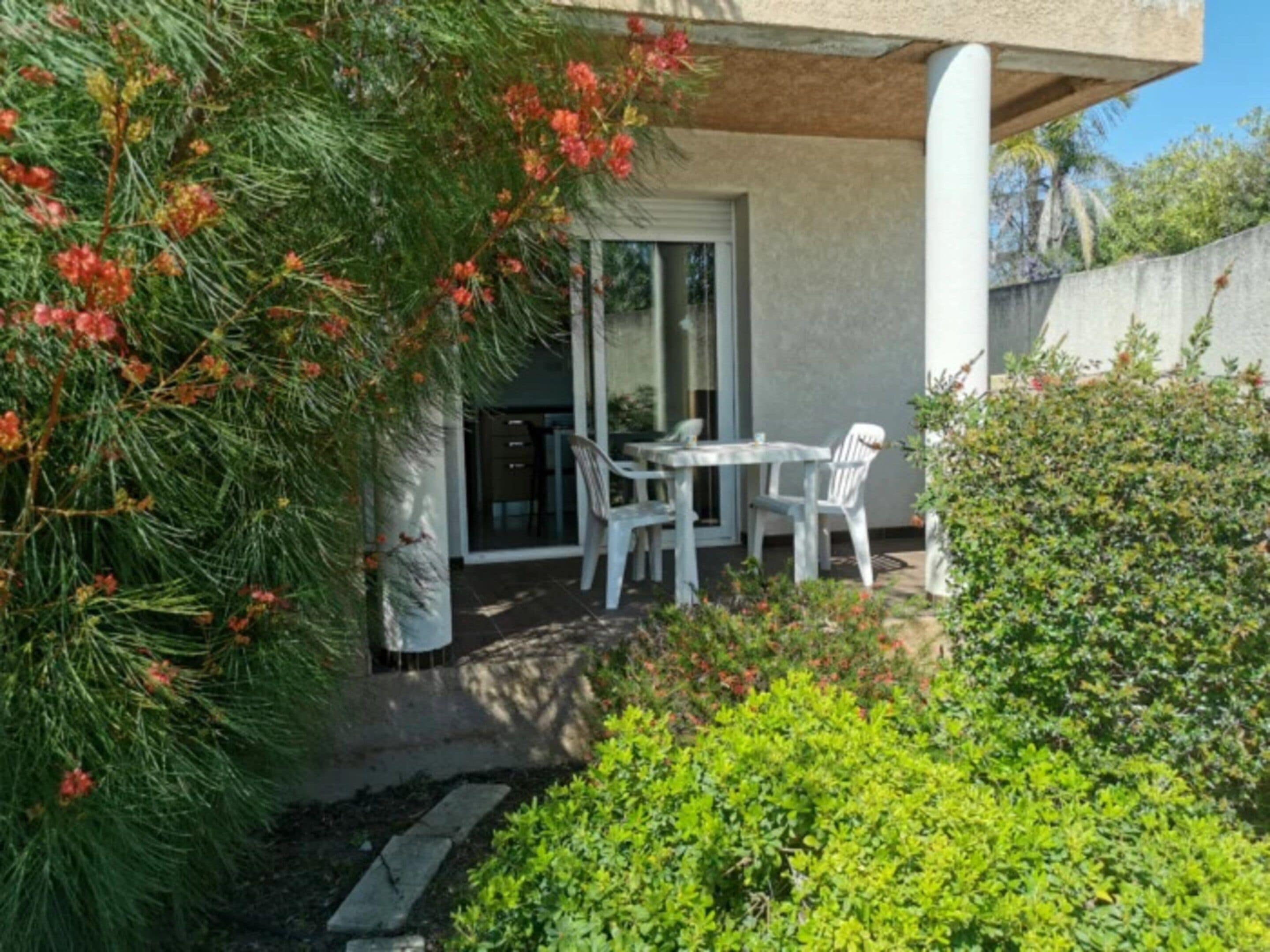 Un patio con una mesa y sillas, una puerta de cristal que da a una habitación y un pequeño jardín con follaje y un árbol de flores rojas.
