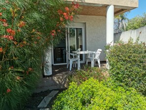 A patio area with a table and chairs, a glass door leading to a room, and a small garden with greenery and a tree with red flowers. - Family Duplex With Terrace in Linguizzetta (Linguizzetta)