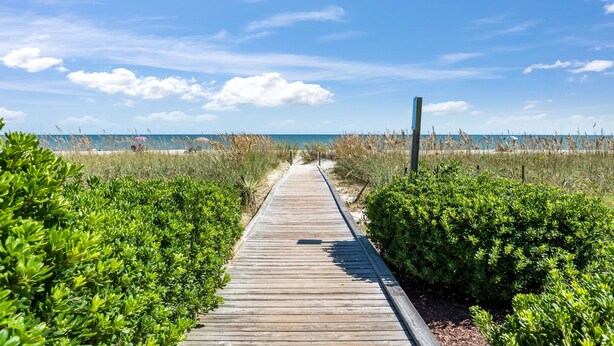 Ein hölzerner Steg führt zu einem Strand mit blauem Himmel und weiter Ferne.