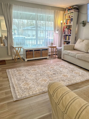 A living room with a sofa, a bookshelf, a window with curtains, a small table, and a patterned rug.