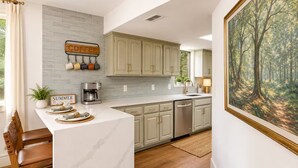 A kitchen with a coffee station, a dining area with a chair, and a painting on the wall.