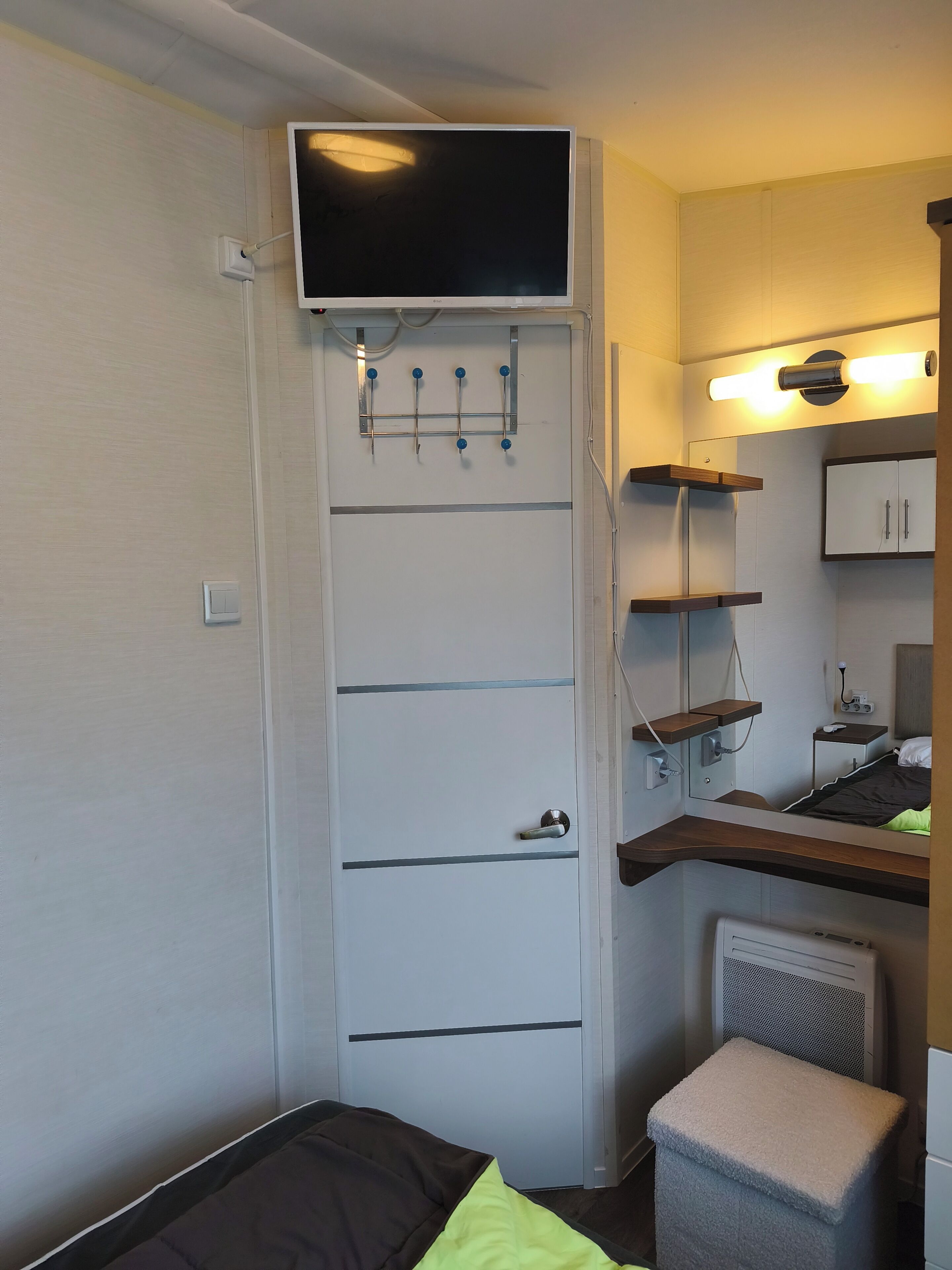 A compact room with a bed, a TV mounted on a wall, a small stool, and a radiator.
