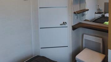 A compact room with a bed, a TV mounted on a wall, a small stool, and a radiator.