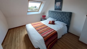 Ein Schlafzimmer mit schrägem Dach, einem Oberlicht, einem Bett mit Steppdecke, einem Nachttisch, einer Lampe und einem gerahmten Bild an der Wand.