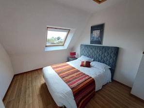 Ein Schlafzimmer mit schrägem Dach, einem Oberlicht, einem Bett mit Steppdecke, einem Nachttisch, einer Lampe und einem gerahmten Bild an der Wand.