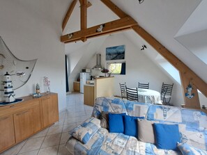 Ein gemütliches Schlafzimmer im Dachgeschoss mit schrägen Wänden, einem Holzbalken an der Decke, einem Bett mit gemusterten Textilien, einer kleinen Küchenzeile und einer Essecke mit Stühlen.
