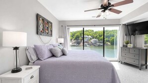 Ein Schlafzimmer mit Bett, Nachttisch, Schminktisch, Deckenventilator und einem großen Fenster mit Blick auf einen See und Gebäude.