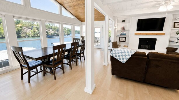 A spacious living area with a dining table, brown leather couch, and a fireplace. Large windows offer a view of a lake. - Lakeside Escape with Boat Slip (Rocky Mount)