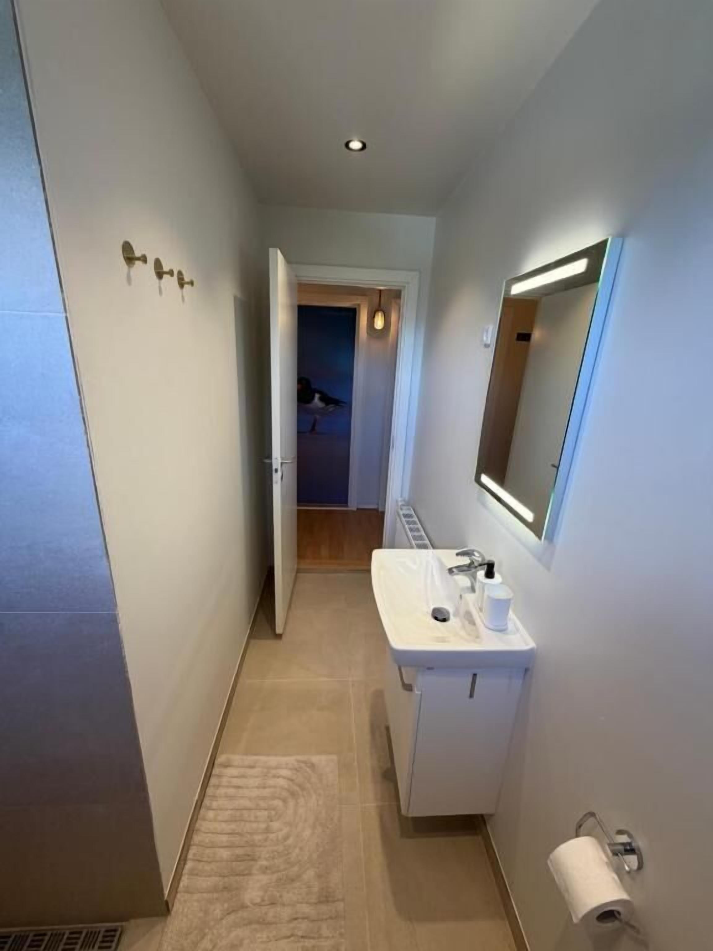 A narrow hallway with a white sink, a mirror, and a door leading to another room.