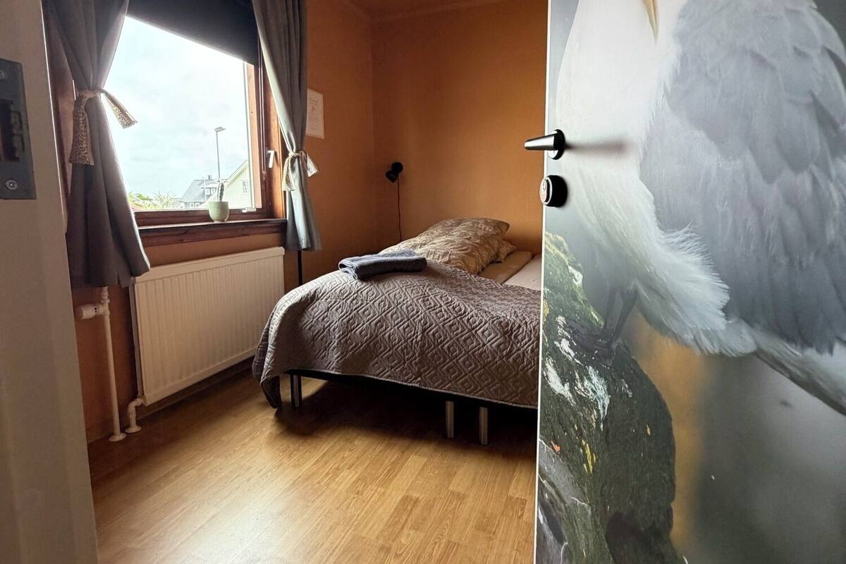 Una habitación con una cama, una ventana con cortinas, un radiador y un cartel de una gaviota en la pared.