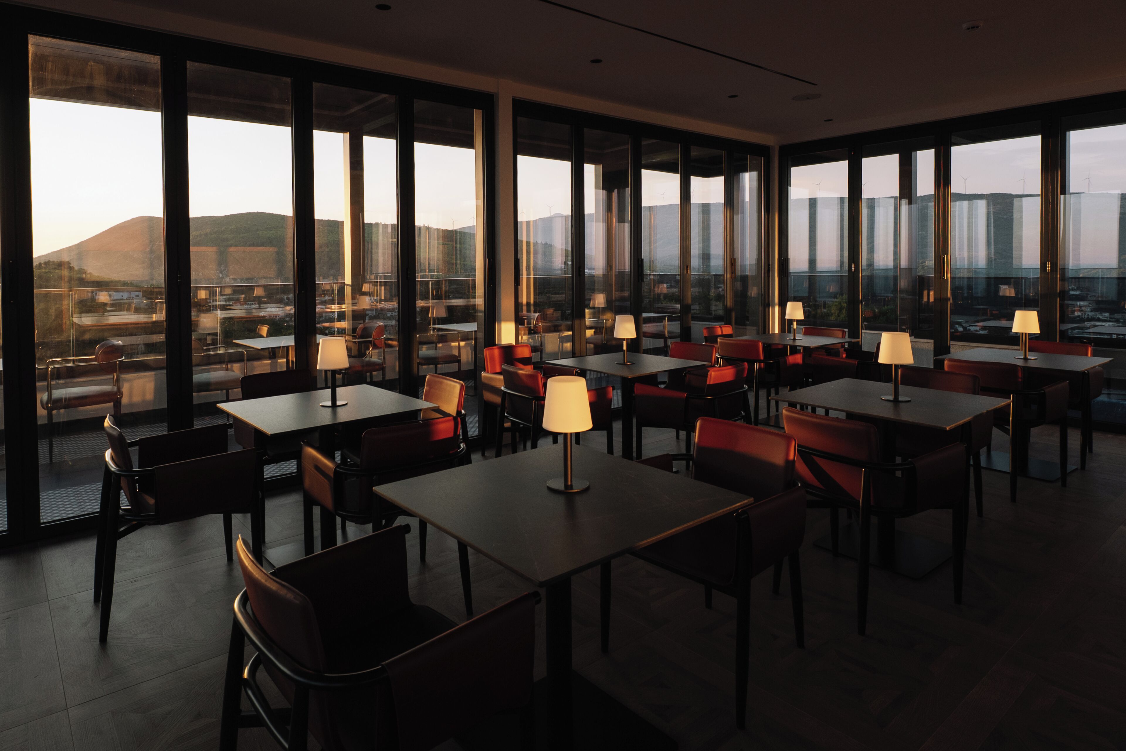 A modern dining area with tables and chairs, large windows offering a view of the cityscape and mountains.