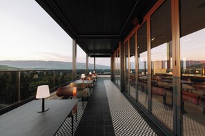 Una terraza con mesas y sillas, ventanas amplias y vistas a aerogeneradores.