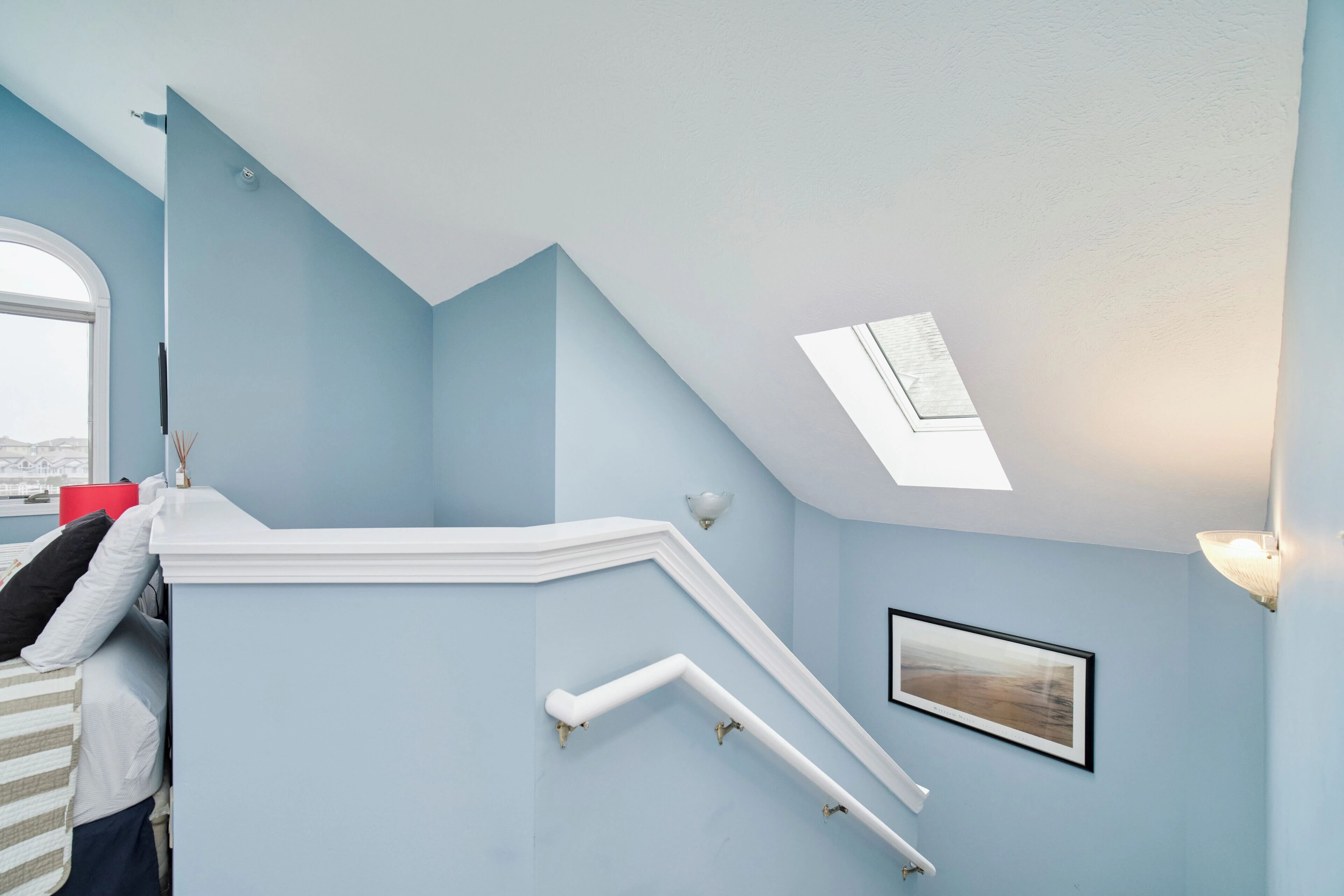 Una habitación con techo inclinado, tragaluz, escalera blanca, un cuadro enmarco y ventana con vistas.