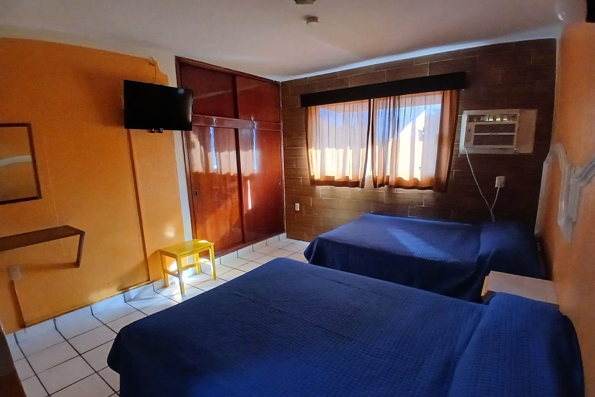 A hotel room with two beds, a TV, a window with curtains, and a brick wall.