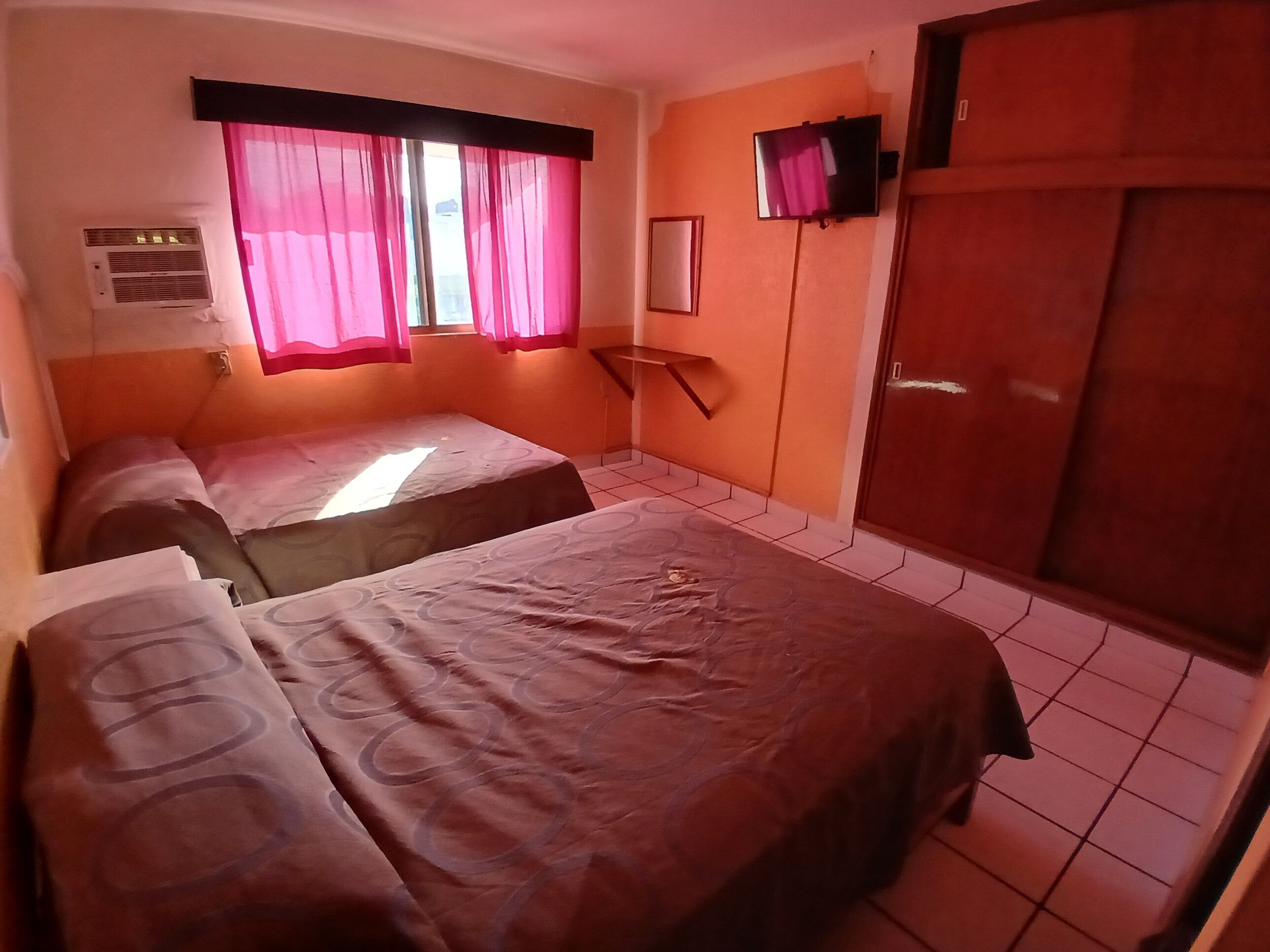 A room with two beds, a TV mounted on the wall, and an air conditioning unit.