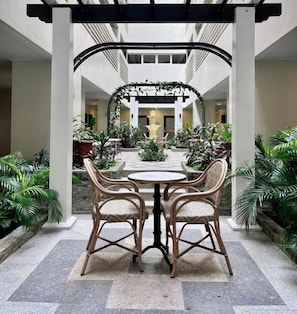 A covered outdoor dining area with a round table and four wicker chairs, surrounded by potted plants and a fountain. - Cozy Studio Living in Iloilo City (Iloilo City)
