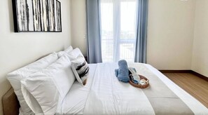 A hotel room with a bed, white bedding, a blue towel, a basket, and a window with curtains. - Relaxing Iloilo Stay with Pool Access (Iloilo City)