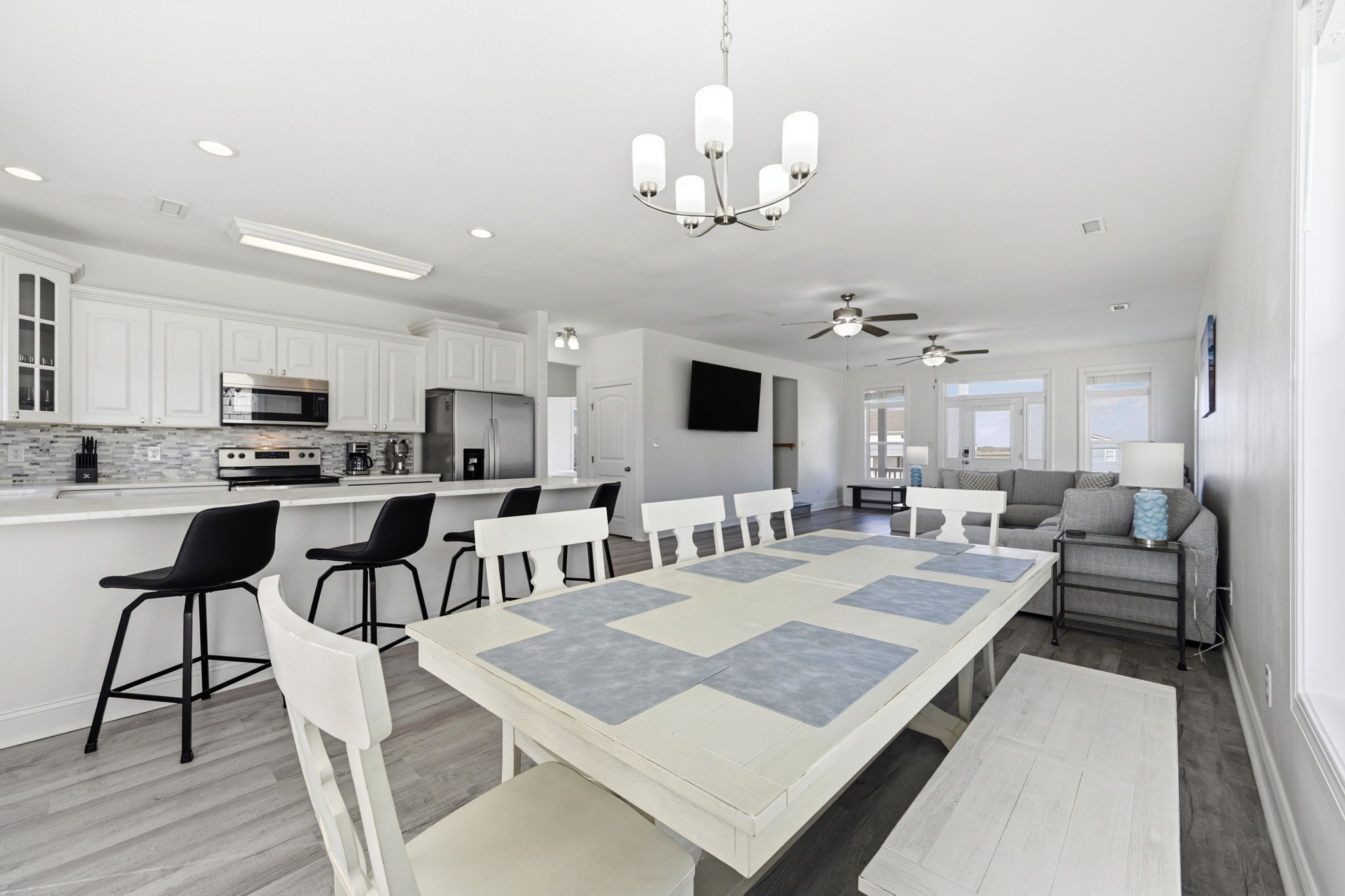 A modern kitchen with white cabinets, a dining area with a large table, and a living area with a sofa and TV.