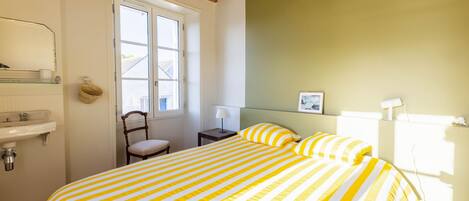 Un dormitorio con cama, ventana, mesita de noche, silla y estante en la pared.