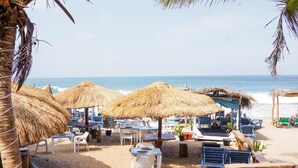 Zona en la playa con sombrillas de paja, mesas y sillas de madera, con vista al océano.