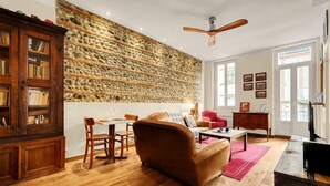 A living room with a stone wall, wooden furniture, a ceiling fan, and a dining area. - L'Halo Gloire - Charmant T2 de caractère (Toulouse)