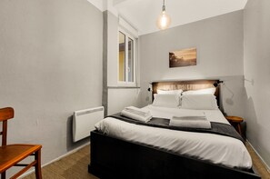 Un dormitorio con una cama, una silla, un radiador, una ventana y un cuadro en la pared.