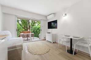 Ein modernes Wohnzimmer mit einer Couch, einem Esstisch mit Stühlen, einem an der Wand montierten Fernseher und einer Glastür, die zu einem Außenbereich mit Tisch und Stühlen führt.