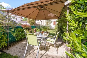 Eine Terrasse im Freien mit Tisch und Stühlen, einem braunen Sonnenschirm und viel Grün.