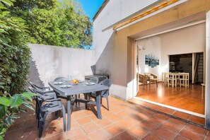 Eine Terrasse im Freien mit Tisch und Stühlen, einem gefliesten Boden und Blick ins Innere durch eine offene Tür.