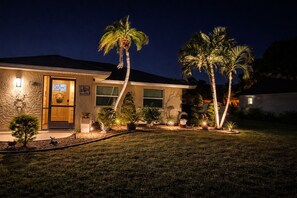 A house with a well-lit entrance, palm trees, and a garden. - The Cozy Coastal Retreat with screened patio and arcades (Sarasota)