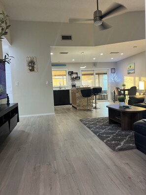 A modern living room with a dark sofa, a coffee table, and a rug. There is a kitchen area with a bar and a dining table. - The Cozy Coastal Retreat with screened patio and arcades (Sarasota)