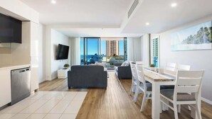 A modern living room with a dining area, a kitchen with a dishwasher, and a large window offering a view of high-rise buildings. - Residence Surfers Ocean and Pool Views (Surfers Paradise)