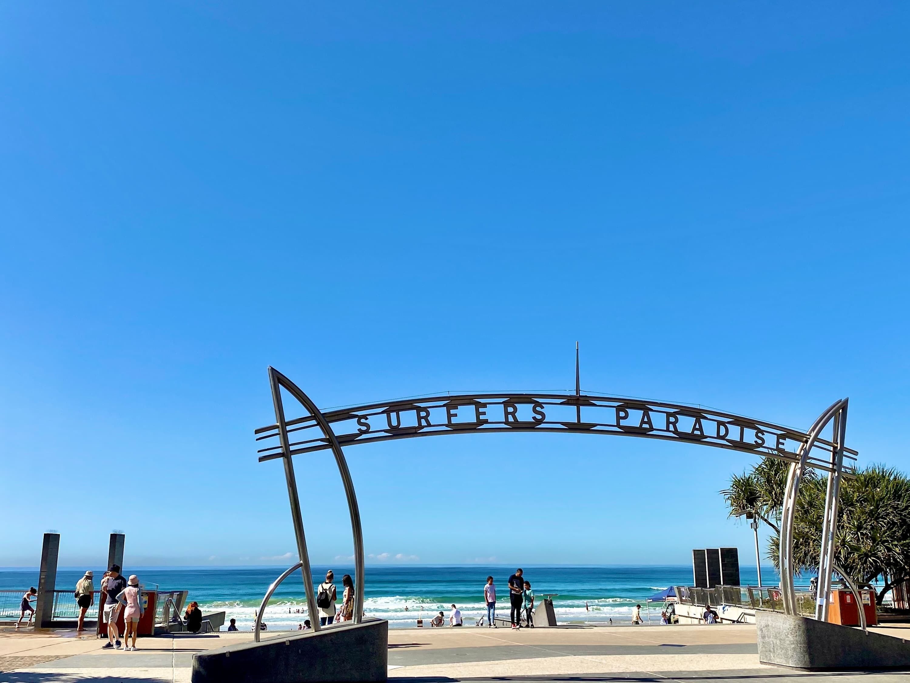 Una entrada a la playa con un arco de metal que dice 'SURFERS PARADISE' y gente caminando por la orilla.