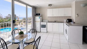 Ett modernt kök med vita skåp, en mikrovågsugn, en ugn och en tvättmaskin. Det finns ett glasbord med stolar och en balkong med utsikt över havet.