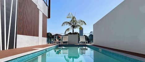 A swimming pool with a clear blue water surface, surrounded by modern buildings with a mix of glass and wood paneling.