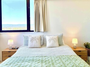 Ein Bett mit einer weiß-grünen Tagesdecke, zwei Kissen mit Palmenstickerei, ein Fenster mit Blick auf eine Stadtlandschaft und zwei Nachttischlampen auf Holznachttischen.