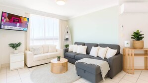 Un salon moderne avec un canapé gris, des fauteuils blancs, une table basse ronde et un téléviseur fixé au mur.