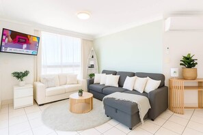 A modern living room with a grey sofa, white armchairs, a round coffee table, and a TV mounted on the wall. - Ocean Getaway 1BR Heated Pool & Netflix (Surfers Paradise)