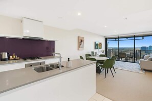 A modern kitchen with a central island, a dining area with green chairs, and a view of the city through large windows. - Resort Retreat in Circle: 2BR Stay & SPA (Surfers Paradise)