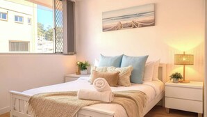 Ein Schlafzimmer mit einem Bett, einem Nachttisch, einer Lampe, einem Gemälde und einem Fenster.