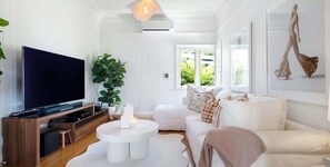 A modern living room with a flat-screen TV, a white sofa, a coffee table, and a large window with a view of greenery.
