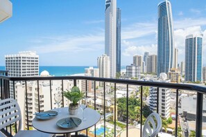 Un balcon avec une table dressée pour le dîner, offrant une vue sur un paysage urbain avec de hauts immeubles et la mer à l’horizon.